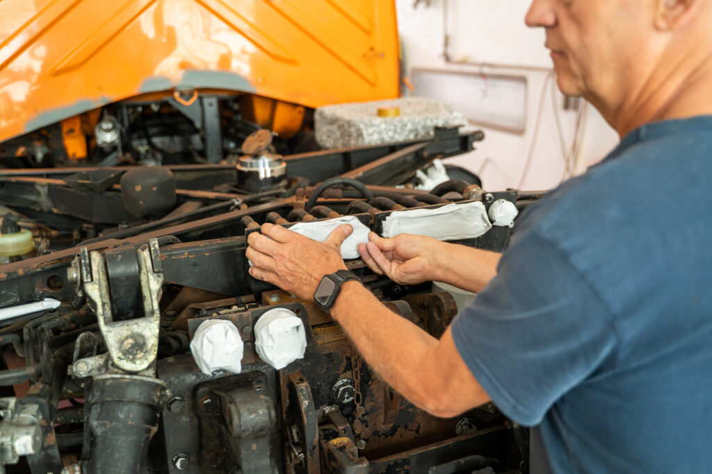 Ein Mitarbeiter klebt Bauteile im Motor- und Fahrwerksbereich eines Fahrzeugs sorgfältig mit Abdeckpapier und Klebeband ab, um sie während der Lackierung vor Farbnebel zu schützen.